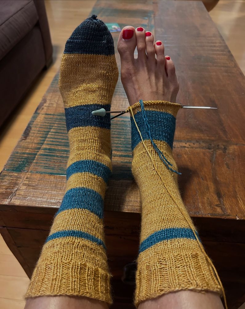 Two feet. One in a sock. One in a partially knit sock. Socks are blue and yellow striped. 
