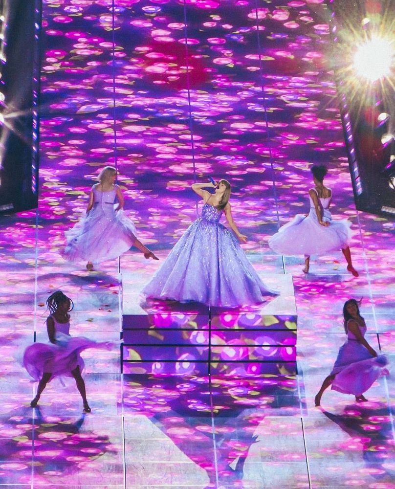 Taylor swift in her speak now purple dress on the speak now eras set singing in the middle of the stage with 4 backup dancers around her