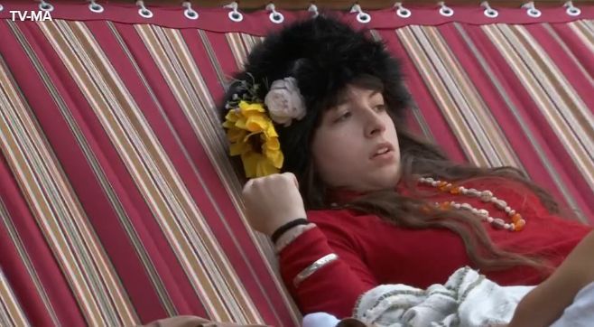 Image grabbed from Hamsterwatch: Ava from BB27 laying in a hammock wearing all red, a big black fluffy hat with flower adornments attached and chunky accessories. She's a tiny very white brunette with a mysterious aura looking very serious. 