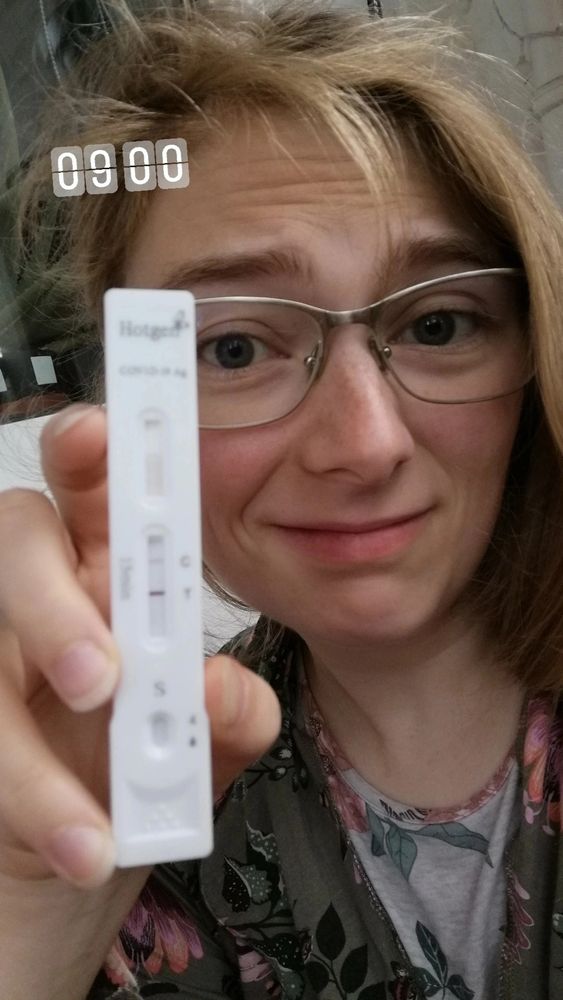 Shari hält einen Coronatest in die Kamera und schaut dabei verzweifelt, aber auch ein bisschen humoristisch. Oben links im Bild steht ein digitaler 09:00h Sticker