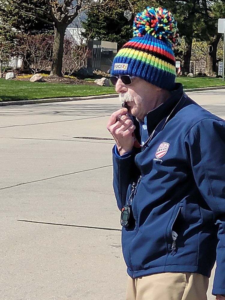A white man with a white soup-strainer mustache l and sunglasses blowing a whistle.
He wears a blue USA Cycling jacket and a blue and multicolor striped winter hat with a giant poofball on top.
