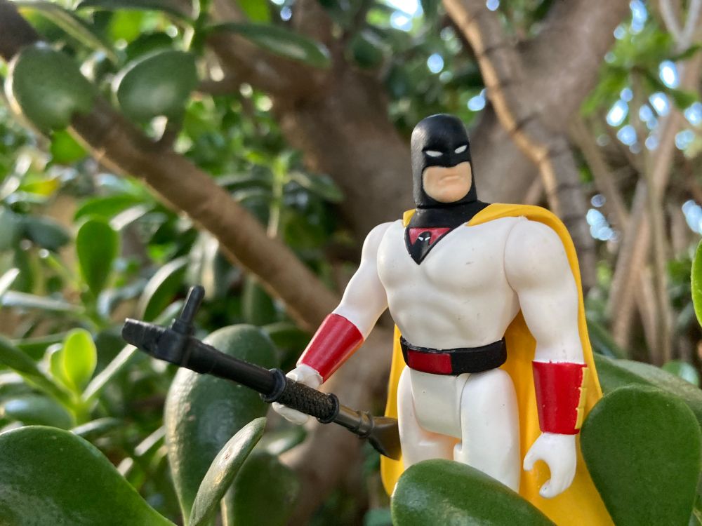An action figure of Space Ghost, a superhero from an old 1960s cartoon, dressed in a white uniforom with black facemask and yellow cape and red bracelets stands amid dark green vegetation holding an iron bar in their right hand. 