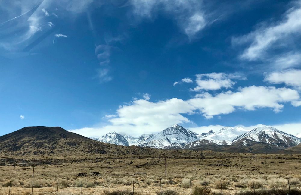 Sierra Nevada from Highway 395. The far away high country and peaks managed by the US Forest Service is snow-covered.  There are high fluffy clouds above the mountains. In the foreground is high-desert range land with sagebrush and dry shrubs 