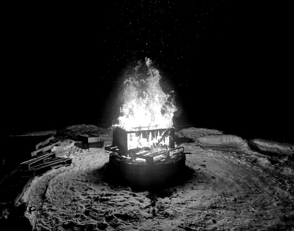 Monochrome photo of a fire in a fire pit during winter time. There is snow on the ground and several wooden pallets are stacked nearby. 
