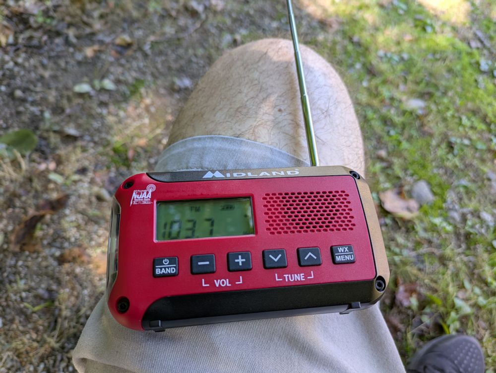 Color photo of a portable and pocket-sized Midland AM FM and weather band radio. The radio is sitting on my leg while sitting outdoors in New Hampshire. The radio is tuned to the FM frequency of 103.7.