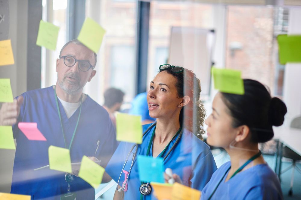 Three healthcare professionals in scrubs collaborate around a glass wall covered in colourful sticky notes.