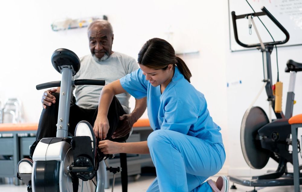 A healthcare worker helps an older adult adjust his foot on a stationary bike in a rehab setting.