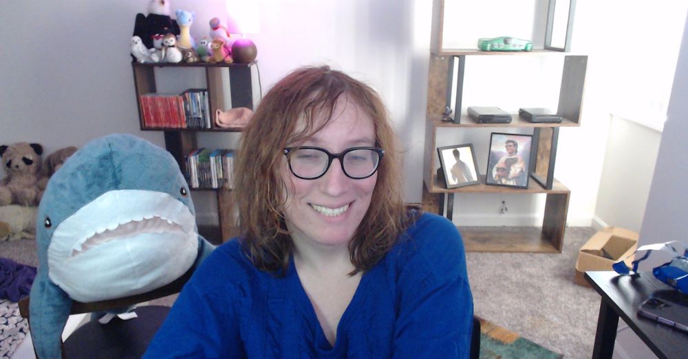 A red-haired woman in a blue top smiles for the camera to announce that she is going live on Twitch! Her trusty stuffed shark Blahaj is at her side.