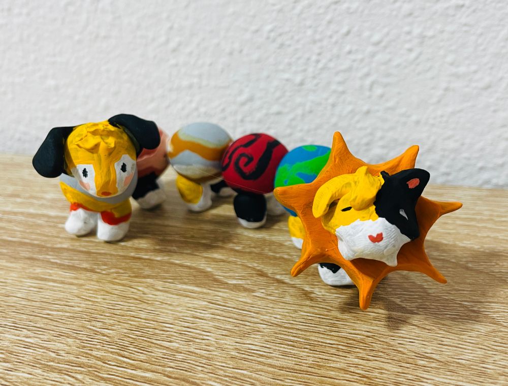 A painted clay figure of a cat and a dog connected by several planets with feet. The cat has sun's rays behind its head like a lion's mane, and the planets are painted in various swirling colors. 