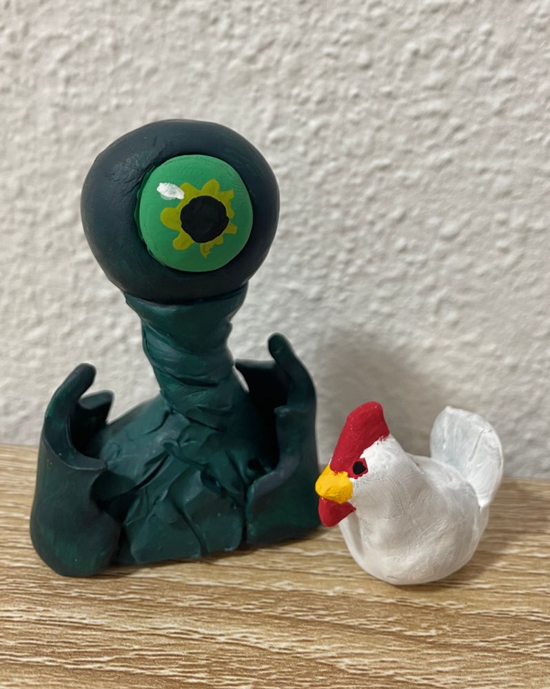 A polymer clay piece of a dark green character with a giant green eyeball for most of its head. It has pointy shoulder pauldrons and a long, twisted neck. Beside him is a small white chicken. 