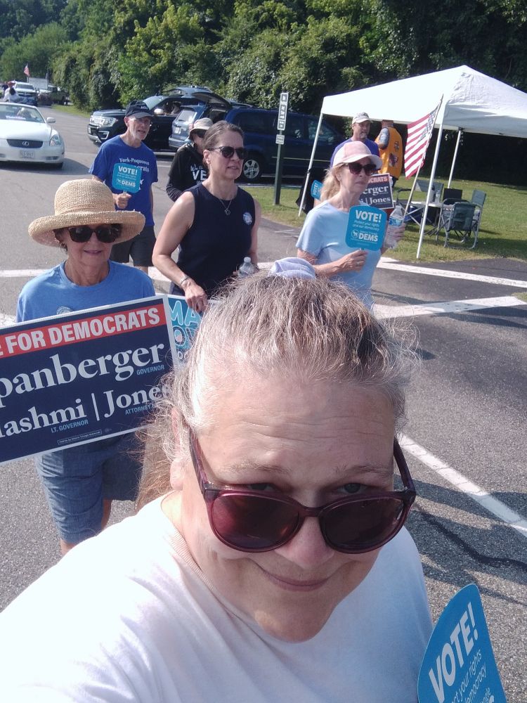 Me grinning and sweating while walking in the Yorktown Independence day parade. Woman  behind me has a nice straw hat and a sign that days "Vote for Democrats: Spanberger for Governor/ Hashmi for Lt. Governor and Jones for Attorney General"