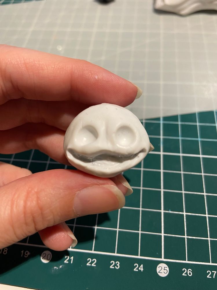 Une petite tête ronde quoique légèrement aplatie avec deux immenses orbites et une large bouche édentée faisant toute la largeur du visage (sculpture en fimo)