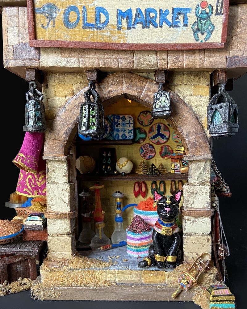 Vue de face Échoppe égyptienne en pierres jaune légèrement ensablée, dotee d’une arche en son centre. Une pancarte au dessus indique « old market ». Plusieurs produits sont visibles de haut en bas : lanternes en laiton, vanneries, bijoux en or, mini pyramides, coffrets à bijoux décorés, narguilés, babouches et épices. Un chat noir Avec un disque et des bracelets d’or se tient calmement devant la boutique 