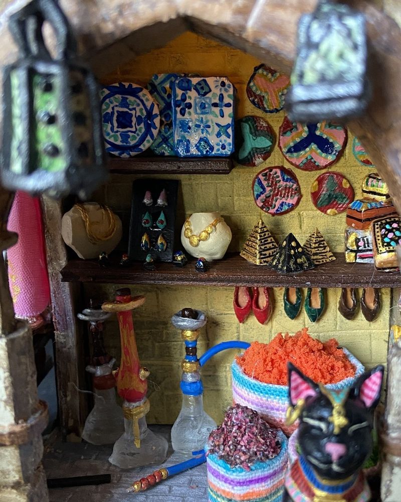 Close up de l’échoppe 3/4 gauche. Plusieurs produits sont visibles de haut en bas : lanternes en laiton, vanneries, bijoux en or, mini pyramides, coffrets à bijoux décorés, narguilés, babouches et épices. Un chat noir Avec un disque et des bracelets d’or se tient calmement devant la boutique 
