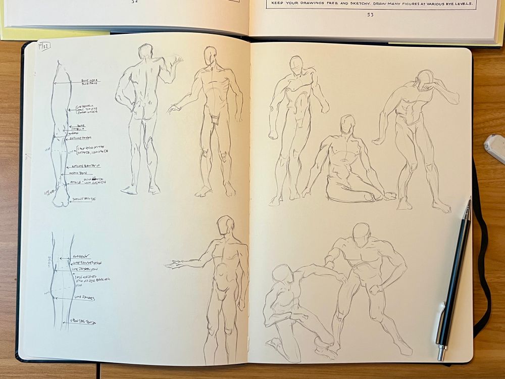 An open sketchbook on a table, the pages are filled with several simplified anatomical sketches of male figures.