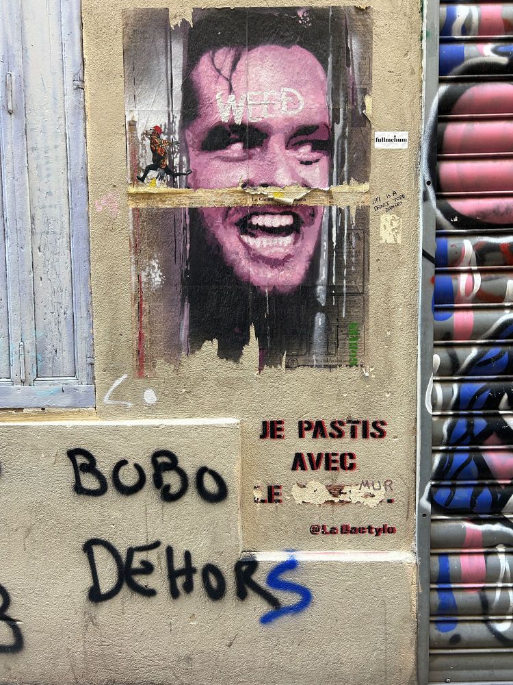 Photo d’un mur de Marseille avec affiche déchirée, collage d’une photo d’homme aux yeux inquiétants (Jack Nicholson dans Shining), tag « Airbnb bobo dehors », pochoir « je pastis avec… »