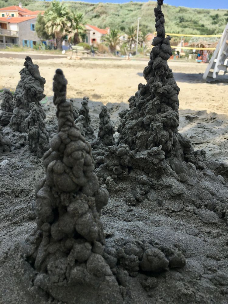 Das Foto zeigt eine Sandburg am Strand, die mit der sogenannten „Tropftechnik“ gebaut wurde. Dabei nimmt man sehr nassen Sand in die Hand und lässt ihn langsam zwischen den Fingern heruntertropfen. Auf diese Weise entstehen die unregelmäßigen, knubbeligen Türme und Spitzen, die man auf dem Bild sieht.
