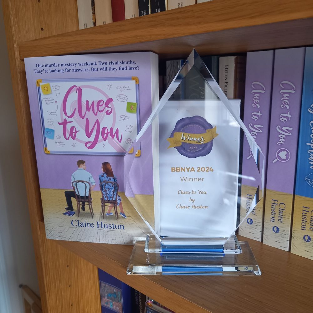 The Book Bloggers' Novel of the Year 2024 winner's trophy for Clues to You by Claire Huston. A crystal, tear-drop shaped trophy on a bookshelf next to a copy of Clues to You.