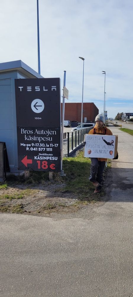 Protestoija seisoo Teslan suuntaviitan vieressä pitäen kylttiä, jossa lukee "Swastikaara: fasistin valinta -- sinunkin?"