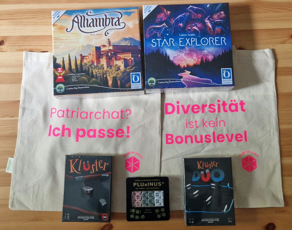 Auf einem Holztisch liegen die Spiele "Alhambra", "Star Explorer", "Kluster", "Kluster Duo" und "PLUxINUS" sowie zwei Jutebeutel mit pinkfarbener Schrift von FLINTA*stisch mit den Sprüchen "Patriarchat? Ich passe!" und "Diversität ist kein Bonuslevel".