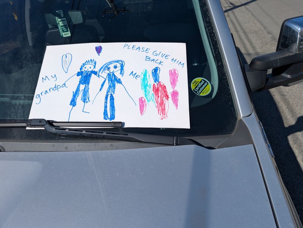 photo of a child's drawing propped up on a front window of a SUV.  there are two figures and a couple amorphous shapes. the figures are labeled "my grandpa" and "me" with a further message over the shapes "please give him back."