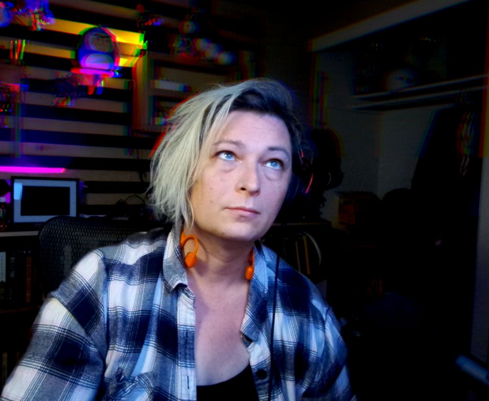 photo of a white woman (@polerin) with chin length blond and dark brown hair, wearing a plaid shirt and two pairs of headphones for some reason.  The background is colorful but distorted for some reason