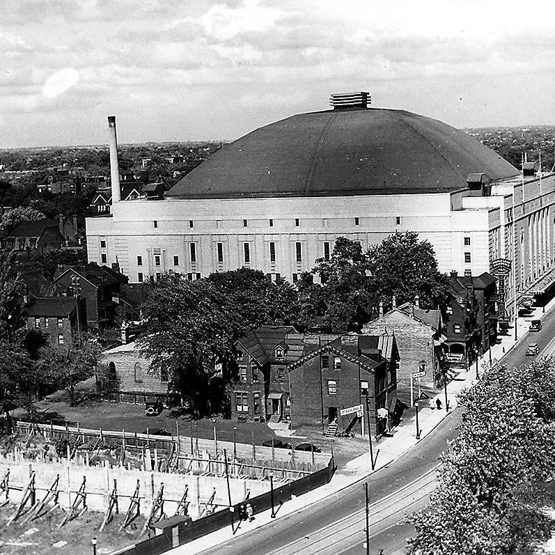 Black and white historical photo from 1939 showing Maple Leaf Gardens in Toronto, a large arena with a prominent domed roof and chimney-like vent on top. In the foreground, a residential street with brick houses, trees, parked vintage cars, and a few pedestrians. The arena's facade has multiple windows and signage. 