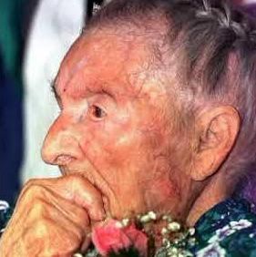 An elderly person with braided hair and wrinkled skin is shown in profile, holding a hand near their face. They are wearing a floral-patterned garment adorned with pink flowers.