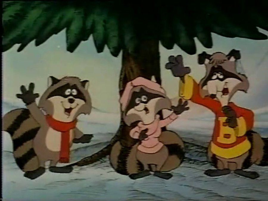 Three animated raccoons standing in the snow under a large evergreen tree. The left raccoon wears a red scarf and waves happily. The middle raccoon, smaller and wearing a pink hooded outfit, raises both arms excitedly. The right raccoon, dressed in a red and yellow jacket, holds a yellow object in one raised hand and waves with the other, mouth open as if singing or cheering. The background shows a snowy landscape with a light blue sky.