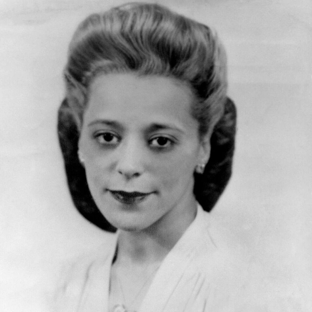 A black-and-white portrait of a woman with neatly styled, voluminous hair, wearing a light-colored dress and a heart-shaped necklace. She is looking directly at the camera with a slight smile.
