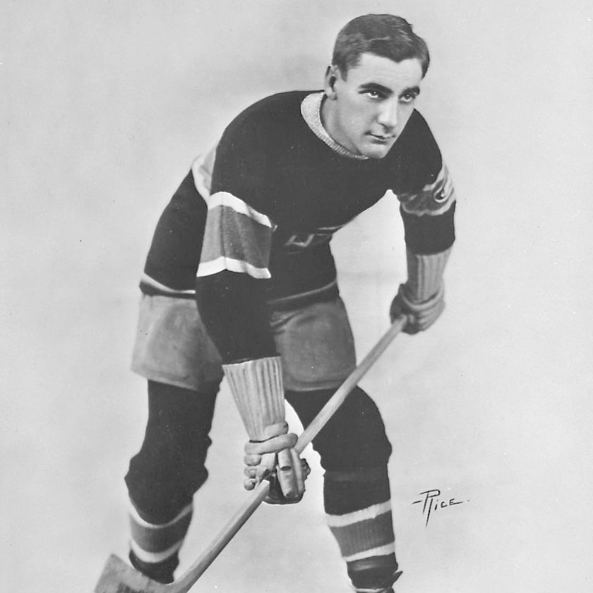 Black and white vintage photograph of ice hockey player Sylvio Mantha posed in a crouched stance on skates, wearing a dark sweater with light stripes on the sleeves and a team logo on the chest, matching shorts, gloves, striped socks, and holding a hockey stick with the blade touching the ice, looking intently downward.