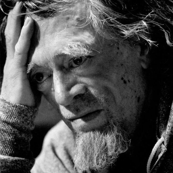 A black-and-white close-up photograph of Milton Acorn with a weathered face, holding his head with one hand. He has a full, white beard and disheveled hair, wearing a textured sweater. The image captures a contemplative or tired expression.