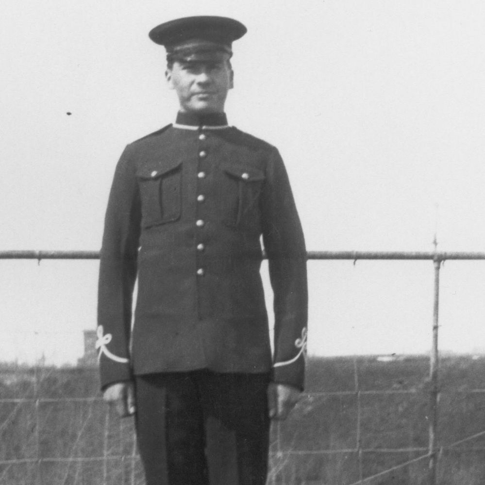 Black-and-white vintage photograph of a man standing outdoors in a uniform from the early 20th century, likely World War I era. He wears a dark wool tunic with high collar, two buttoned chest pockets, braided cuffs, and a peaked cap. His hands are at his sides, posture formal. The background shows an open field with a barbed wire fence and distant landscape, typical of a military training ground or camp. The image has a slightly grainy, aged quality.