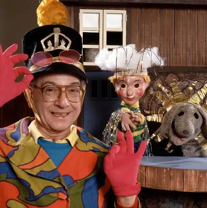 A person wearing a colorful, abstract-patterned outfit and a black hat with a yellow plume and pink gloves is making hand gestures. Behind them, a puppet with a white, fluffy hat and green outfit stands next to a gray dog puppet adorned with gold decorations. The background features a wooden house with white-framed windows and a textured wall with golden accents.