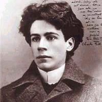 A sepia-toned photograph of a young man with curly dark hair, wearing a high-collared shirt and a double-breasted coat with a pin on the lapel. The background contains handwritten text in an unknown script.