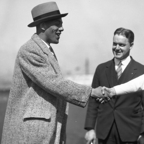 A black-and-white photo showing two men shaking hands outdoors. The man on the left is wearing a light-colored suit and a fedora hat, smiling broadly. The man on the right, also smiling, is dressed in a dark suit with a tie. A third person, partially visible, extends a hand from the right side, wearing a light-colored sleeve. The background is blurred, suggesting an outdoor setting, possibly near a body of water or a ship.
