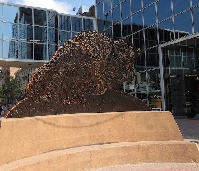 The photo depicts a large, abstract sculpture of a bison's head, likely made of metal or a similar material. The sculpture has a textured, patterned surface with intricate designs, giving it a rugged yet artistic appearance. It is mounted on a curved, beige stone pedestal, which elevates it slightly off the ground. The background features a modern urban setting with tall glass buildings, reflecting the sky and surroundings. The sky is partly cloudy, and there are some trees and people visible in the distance, suggesting this is a public space, possibly a plaza or courtyard in a city. The overall scene combines natural elements (the bison sculpture) with a contemporary architectural environment.