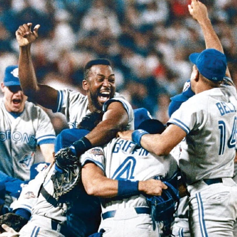 A jubilant group of Toronto Blue Jays baseball players celebrating a victory, with one player being lifted and cheered by teammates wearing gray uniforms with blue accents and caps, in front of a large, excited crowd in the stadium.