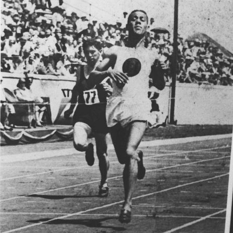 A black-and-white historical photo of two athletes racing on a track, with one wearing a numbered jersey (77) and the other in a darker uniform. The scene is set in a stadium with a large crowd of spectators in the background, seated in stands. The track is marked with lanes, and a pole is visible on the right side. The photo is credited to an unknown source from the Wolf Reinhart Archive (cropped).