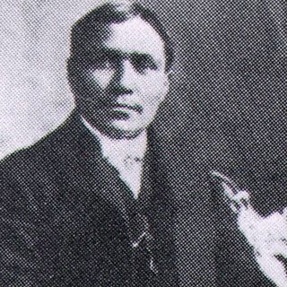 A black-and-white photograph of a man wearing a dark suit and tie, holding a cigarette in one hand, with a textured background.