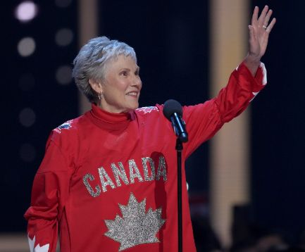 The photo shows Anne Murray wearing a vibrant red outfit with the word "CANADA" and a large maple leaf design on the front, suggesting a patriotic theme. She has short, gray hair and is raising her right hand in a wave. She is standing in front of a microphone, indicating she might be performing or speaking on stage. The background is blurred with soft lights, typical of a concert or event setting.