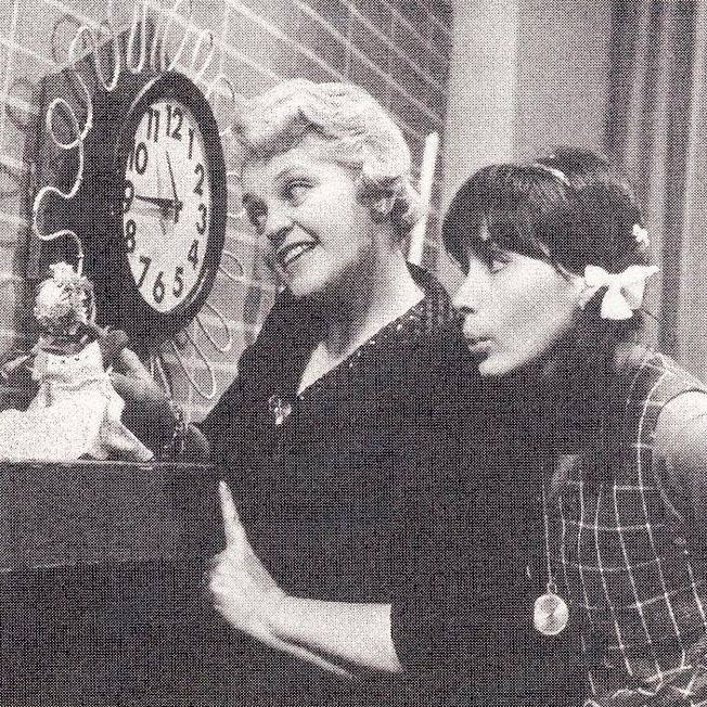Two people, one older woman and one younger girl, standing near a clock on a wall. The older woman is pointing with a smile, while the younger girl is making a playful face. A small doll or figurine is placed on a surface below the clock. The scene appears to be indoors with a brick wall and curtain in the background.