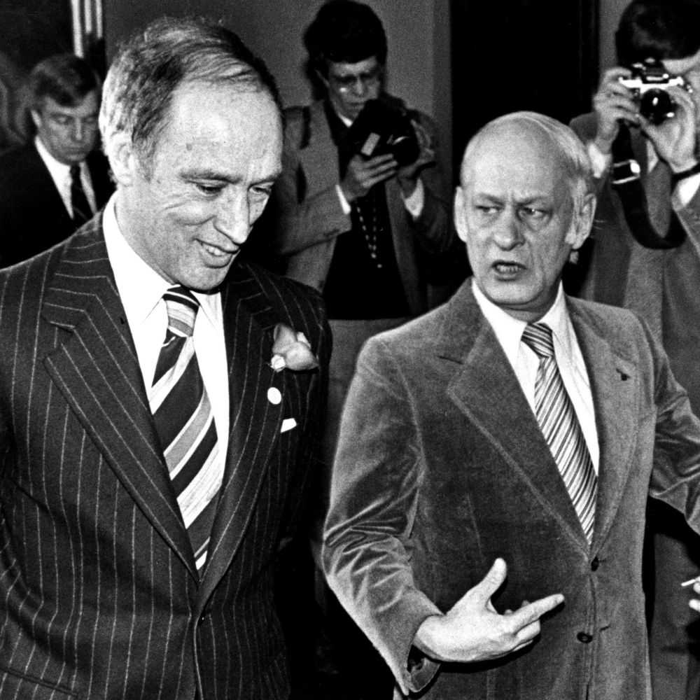 Black and white photo of Pierre Trudeau and René Lévesque, both men with white hair, wearing suits, standing together at a public event, surrounded by a crowd and photographers, with microphones and banners in the background.