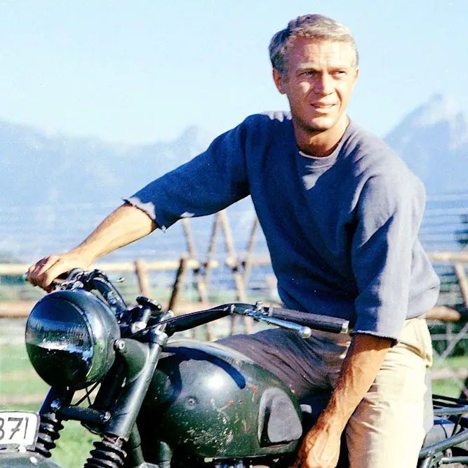A middle-aged man with short, neatly combed gray hair, piercing blue eyes, and a slight smile rides a vintage olive-green motorcycle in a sunny, open landscape. He wears a loose-fitting navy blue sweatshirt with the sleeves rolled up, khaki chinos, and brown boots, leaning casually on the handlebars with his right hand. The motorcycle has a round headlight, black leather seat, and a license plate reading "WH-13371." In the background, wooden corrals and fences frame a grassy field under a clear blue sky, with distant snow-capped mountains (resembling the Tetons) on the horizon.