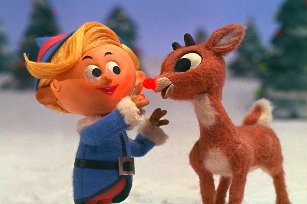 This image is from the classic Christmas television special "Rudolph the Red-Nosed Reindeer" (1964). It features two of the main characters:

Hermey the Elf: On the left, Hermey is depicted with his blonde hair and wearing his blue and white outfit, which is typical for an elf in the North Pole setting. Hermey is known for wanting to be a dentist rather than making toys like the other elves.
Rudolph the Red-Nosed Reindeer: On the right, Rudolph is the young reindeer with the distinctive shiny red nose, which is a central element of the story. He is shown here with his large eyes and brown fur.

In this scene, Hermey appears to be inspecting or talking about Rudolph's red nose, highlighting a moment of friendship and support between the two characters who both feel like outcasts in their respective communities. The background shows a snowy landscape with Christmas trees, reinforcing the winter holiday theme. 