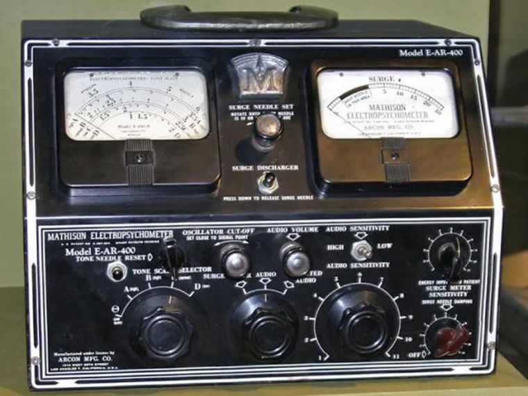The photo shows a vintage electronic device, labeled "Mathison Electropsychometer Model E-AR-400," manufactured by Arcon Mfg. Co. It features a black metal casing with two analog meters at the top, one marked "Tone Needle" with a scale and the other labeled "Surge" with a similar scale. The front panel includes several black knobs for adjusting settings such as "Tone Selector," "Oscillator Cutoff," "Audio Volume," "Audio Sensitivity," and "Surge Meter Sensitivity." There is also a "Surge Discharger" button between the meters. The device is placed on a surface, possibly for display.