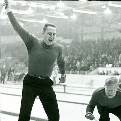 Black-and-white photo of a curling rink: a skip in a dark turtleneck stands in the house with his right arm raised high and mouth open, intensely shouting instructions. In the foreground, his teammate is crouched low in the hack, gripping the broom and preparing to deliver the stone toward the rings where several rocks are already placed. A packed crowd watches from the stands in the background.