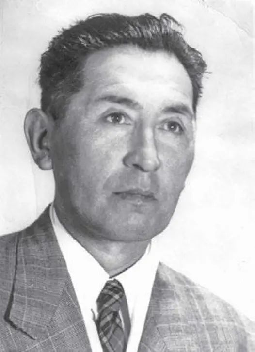 The photo is a black-and-white portrait of a man. He has short, dark hair and is wearing a checkered suit jacket, a white dress shirt, and a patterned tie. The background is plain and light-colored, keeping the focus on the subject.