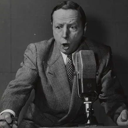 A black-and-white photo of a man in a suit and tie, speaking passionately into a vintage microphone at a desk, with a dark background.