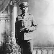 The black-and-white photograph depicts a person standing in a formal pose, dressed in a military uniform. The uniform includes a cap, a buttoned jacket with pockets, trousers, and sturdy boots. The background features a painted studio setting with columns, draped fabric, and decorative elements, typical of early 20th-century portrait photography.